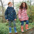 Two children wearing Muddy Puddles jackets and trousers are stood on a fallen tree branch in a wooded area. One wears a 'Blizzard Kids Ski Jacket Pink'.